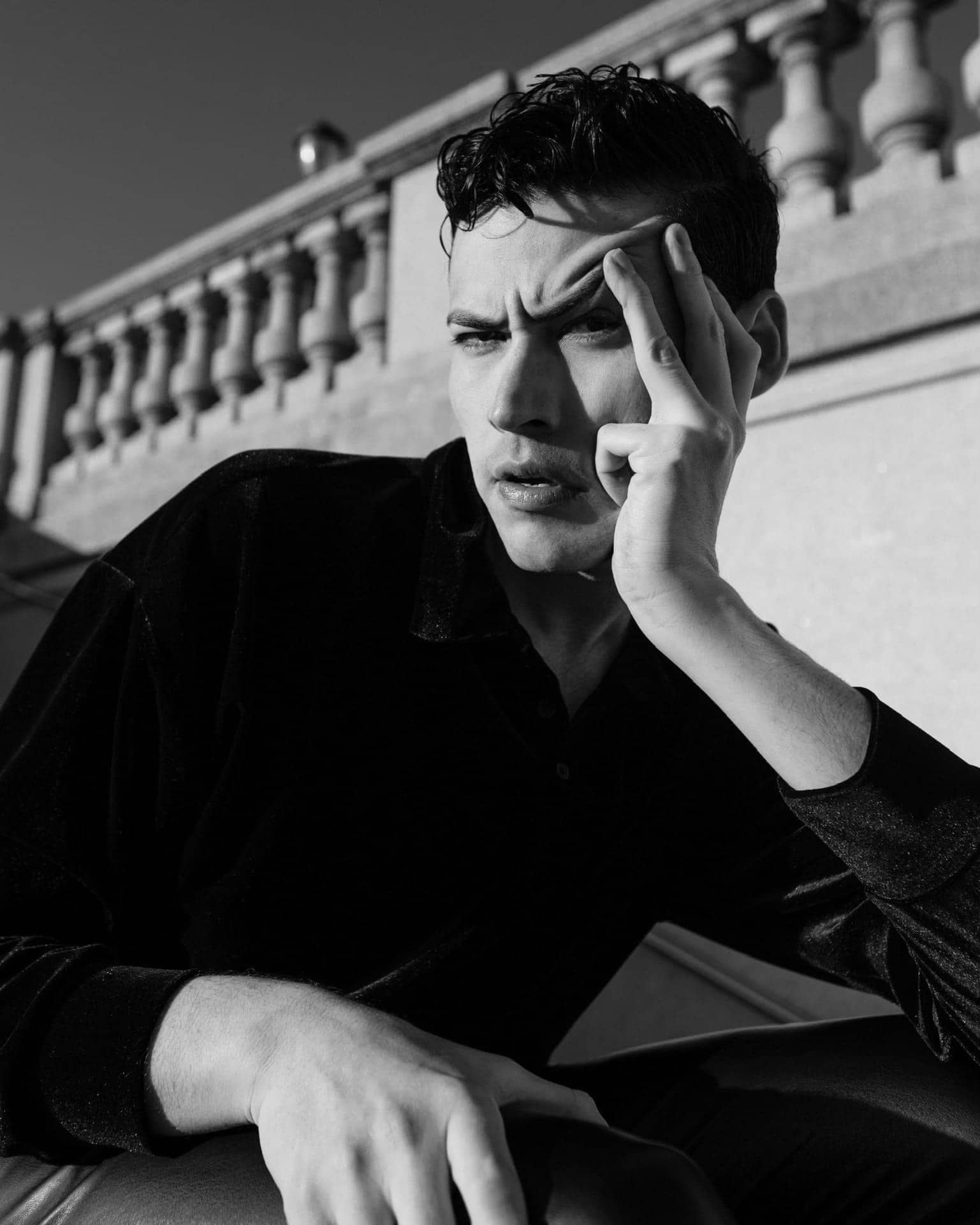 Professional black and white IMDB profile picture of rising star Actor Troy Bronson, striking a unique pose with his left hand on his face, wearing a long-sleeve black shirt and leather pants.