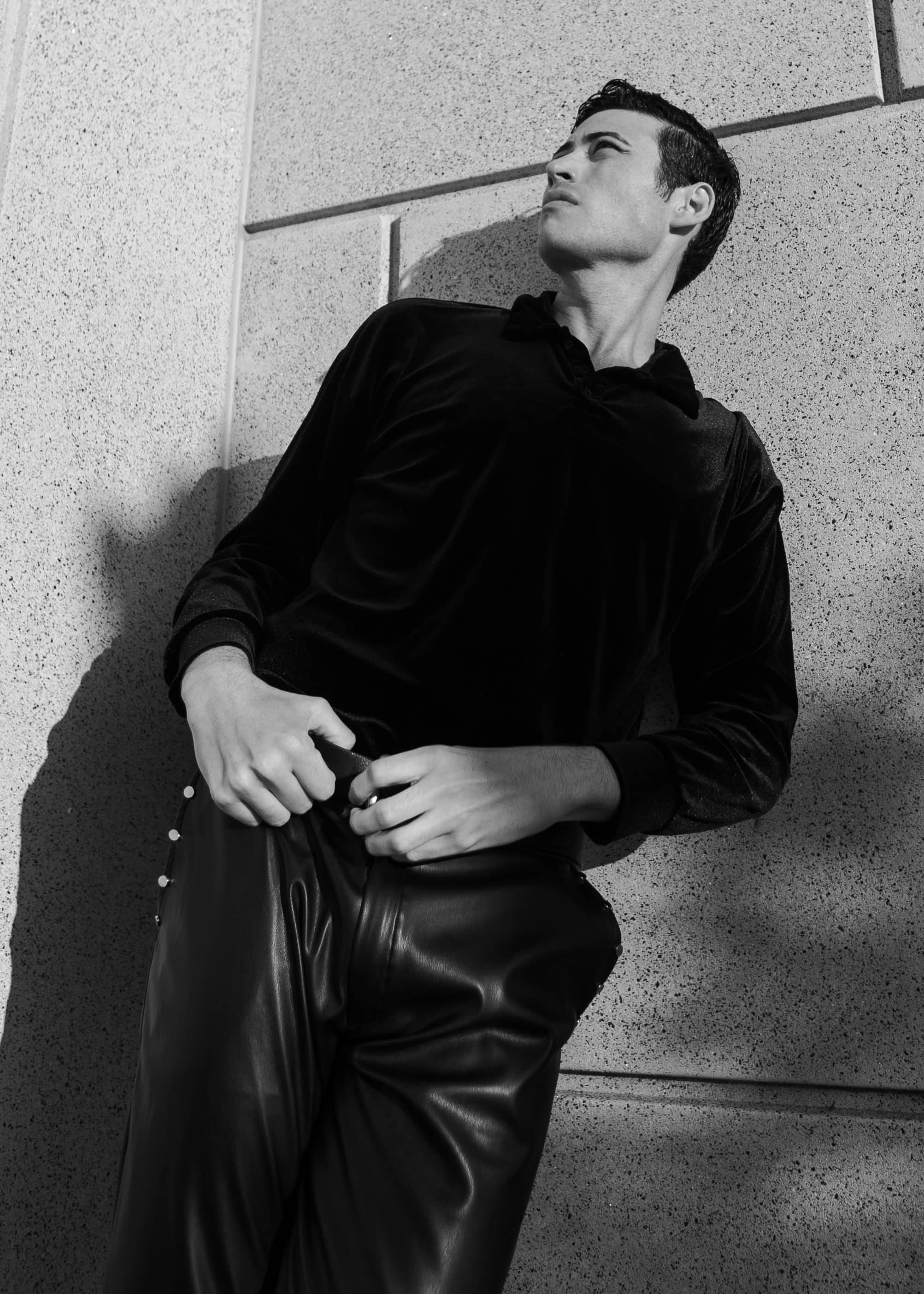 Actor Troy Bronson looking up to the right in a black and white photo, displaying a defined jaw, sleek haircut, and a pose reminiscent of the Hollywood golden age of cinema. He's wearing black leather pants and a velvet long-sleeve shirt, his thumbs hooked into his belt loops, giving an appearance similar to a sculpture.