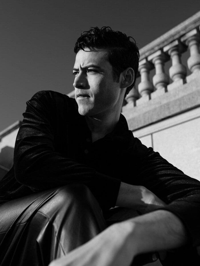 Black and white portrait of Troy Bronson sitting in front of a classical stone balustrade, gazing into the distance with an intense, thoughtful expression, subtly biting his lower lip. He’s dressed in a dark textured shirt and leather pants, with shadows playing across his face and arms, capturing a cinematic moment of introspection and silent determination.